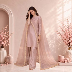 May include: A monochromatic dusty rose ensemble consisting of a long-sleeved tunic and matching wide-leg trousers in a smooth, satin-like fabric. A sheer, floor-length organza dupatta with intricate gold floral embroidery along the edges.