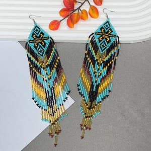 May include: A pair of beaded earrings with a turquoise, black, yellow, and maroon color scheme. The earrings have a diamond-shaped top with a geometric design and long fringe. The earrings are made with small beads and have silver hooks.