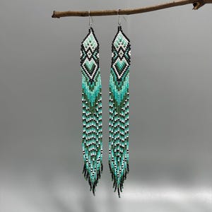 May include: Long beaded earrings in shades of turquoise, black, white, and green. The earrings are suspended from a branch, showcasing a geometric pattern at the top and a fringed bottom. The earrings are approximately 6 inches long.
