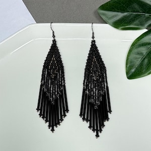 May include: A pair of black beaded earrings with a long, fringed design. The earrings feature a triangular shape with a beaded top and long, cylindrical beads at the bottom. The earrings are displayed on a white surface.