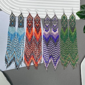 May include: Six pairs of beaded dangle earrings in various colors. The earrings feature intricate geometric patterns at the top and long, flowing beaded fringe. Colors include blue, orange, purple, and green. Each earring has a silver-tone hook.