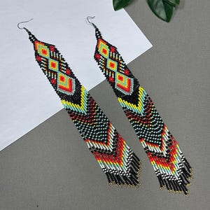 May include: A pair of long, beaded earrings with a geometric design. The earrings feature a black background with red, yellow, and turquoise accents. The bottom of the earrings have a fringe of black and gold beads. The earrings are approximately 6 inches long.