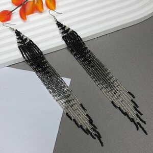 May include: A pair of long, beaded earrings with a gradient design. The earrings transition from black at the top to silver at the bottom, with a fringe-like effect. The earrings are on a gray surface.