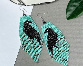May include: Handmade beaded earrings with a black raven design on a turquoise background. The earrings have a fringe at the bottom and silver-coloured ear wires. A unique accessory.