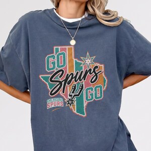 May include: A slate blue short-sleeve t-shirt featuring a graphic of the state of Texas in muted stripes. The design includes the text "GO Spurs GO" in black and white, the San Antonio Spurs logo, and a star icon. The shirt has a crew neckline.