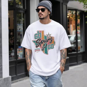 May include: A white short-sleeved t-shirt featuring a graphic of the state of Texas in muted green, pink, and orange tones. The text reads GO Spurs GO with the San Antonio Spurs logo and a star. The design includes the text SAN ANTONIO SPURS.