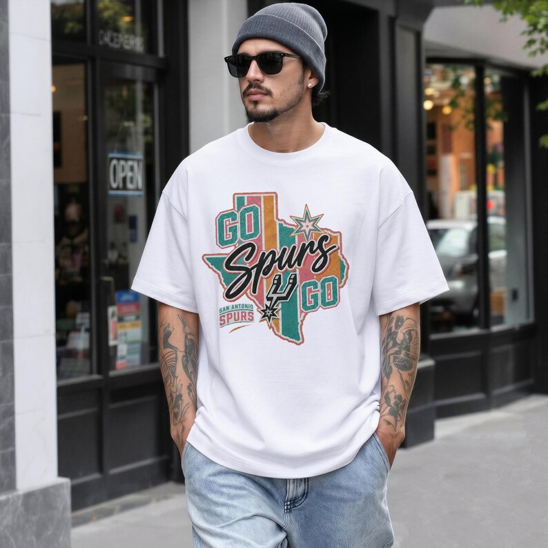 May include: A white short-sleeved t-shirt featuring a graphic of the state of Texas in muted green, pink, and orange tones. The text reads GO Spurs GO with the San Antonio Spurs logo and a star. The design includes the text SAN ANTONIO SPURS.