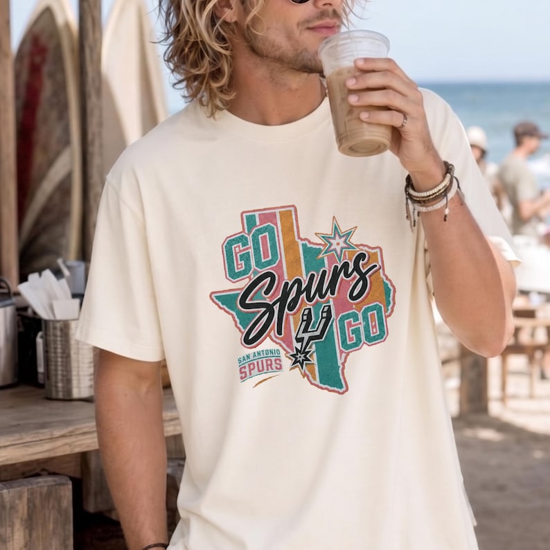 May include: A cream-colored short-sleeve t-shirt featuring a graphic of the state of Texas in muted teal, pink, and orange stripes. The text reads "GO Spurs GO" in black script and "SAN ANTONIO SPURS" with the team logo and a star icon.