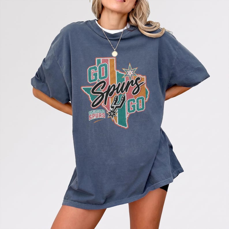 May include: A slate blue short-sleeve t-shirt featuring a graphic of the state of Texas in muted stripes. The design includes the text "GO Spurs GO" in black and white, the San Antonio Spurs logo, and a star icon. The shirt has a crew neckline.