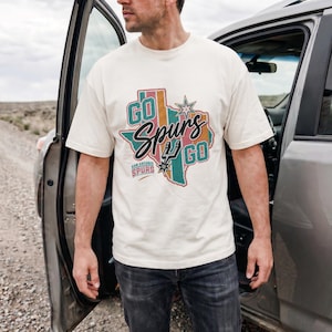 May include: A cream-colored short-sleeve t-shirt featuring a graphic of the state of Texas in muted teal, pink, and orange stripes. The design includes the text "GO SPURS GO" and "SAN ANTONIO SPURS" with a star and the team logo in the center.