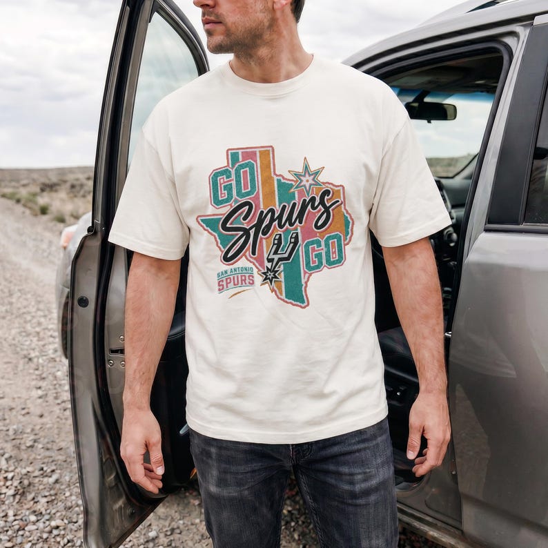 May include: A cream-colored short-sleeve t-shirt featuring a graphic of the state of Texas in muted teal, pink, and orange stripes. The design includes the text "GO SPURS GO" and "SAN ANTONIO SPURS" with a star and the team logo in the center.