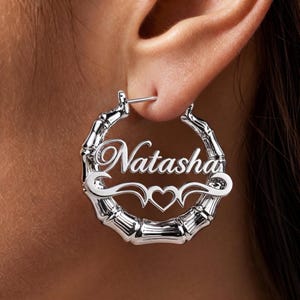 Op de afbeelding: Een zilverkleurige oorring met een textuur die is geïnspireerd op bamboe. In het midden van de ring staat de naam Natasha in een cursief lettertype, geplaatst boven een decoratieve versiering die in het midden een kleine hartvorm bevat.
