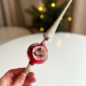 May include: A vintage Christmas tree topper. The red, round ornament has a floral design and a silver spire. The base is silver. The topper is held by a hand.