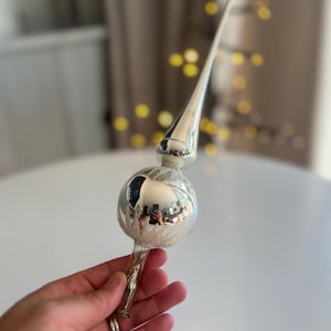 May include: A silver glass Christmas tree topper featuring a central spherical ornament topped with a long, slender, pointed spire. The base of the sphere is decorated with white, textured, snow-like glitter accents that extend upward.