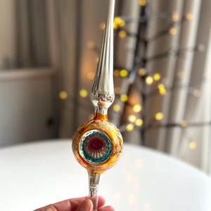 Può includere: Un puntale per albero di Natale in vetro in stile vintage, caratterizzato da una lunga guglia argentata sopra una sfera rotonda color ambra. Il centro della sfera presenta un incavo con motivo a stella nei colori turchese e magenta.