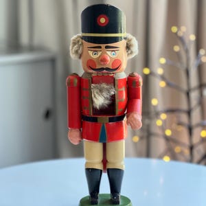 Può includere: Una statuetta schiaccianoci in legno che indossa un cappello nero con un cerchio rosso, una giacca rossa con finiture dorate e stivali neri. La figura ha un volto dipinto con baffi, guance rosate e un ciuffo di pelliccia sintetica sul petto.