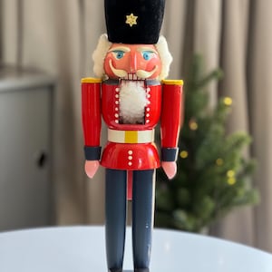 May include: A wooden nutcracker figurine wearing a tall black velvet hat with a gold star emblem, a bright red jacket with white trim, a white faux fur beard, and dark blue trousers. The figure stands on a round gray base with painted details.