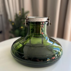 Könnte beinhalten: Ein dunkelgrüner Glasschirm mit breitem, abgerundetem Boden und verjüngtem Hals. Am oberen Ende befindet sich ein Metallkragen mit der Prägung BULACH am Rand, der durch kleine Metallklammern an den Seiten befestigt ist. Das Glas ist glatt und glänzend.