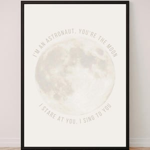 May include: A framed wall art print featuring a large, detailed image of the moon centered on a light cream background. The text "I'M AN ASTRONAUT, YOU'RE THE MOON" arches over the top, and "I STARE AT YOU, I SING TO YOU" arches below.