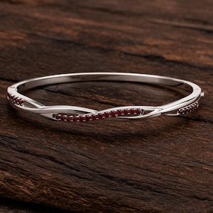 Può includere: Un bracciale rigido incernierato color argento caratterizzato da un design intrecciato e incrociato. La parte anteriore del bracciale è intarsiata con una fila di piccole pietre preziose rotonde rosso scuro che seguono la curva della struttura.
