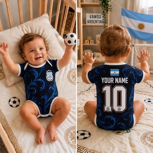 Puede incluir: Un body para bebé de color azul oscuro con un patrón de remolinos, ribetes blancos y el escudo de la selección nacional de fútbol de Argentina en el frente. La parte trasera muestra el texto 'YOUR NAME', el número '10' y una bandera argentina.