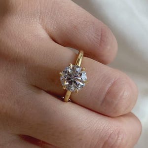 May include: A solitaire ring featuring a large, round-cut, clear gemstone set in a polished yellow gold band. The stone is secured by six prongs, creating a classic look with a high-shine finish that reflects light across its faceted surface.