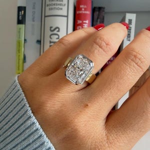 May include: A large rectangular-cut diamond ring with a gold band and a silver-colored bezel. The ring is on a hand with red nail polish, with a light blue sweater sleeve in the background. Bookshelves are visible in the background.