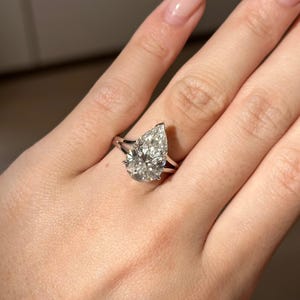 Op de afbeelding: Een peervormige diamanten verlovingsring met een split-shank band. De heldere diamant schittert, gezet in een zilveren of witgouden band. De ring wordt op een hand getoond.