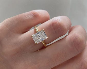 Puede incluir: Un anillo de oro con una gran piedra preciosa transparente de corte radiante rectangular, sujeta por cuatro garras. La banda es de metal de oro amarillo pulido, liso y sencillo. La piedra está facetada y refleja la luz.