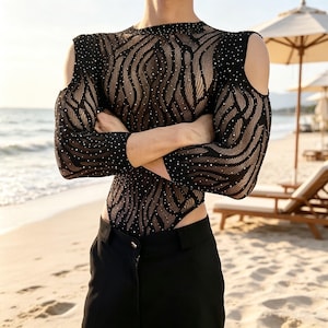 Peut inclure: Un body noir à manches longues en tissu maille transparent avec un motif ondulé texturé et des strass argentés éparpillés. Le vêtement présente des découpes aux épaules, une encolure ronde et une échancrure haute, porté avec un pantalon noir.