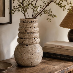 May include: A tall, cylindrical ceramic vase with a porous, speckled beige and brown surface. The vase features a wide, rounded base that tapers upwards into a series of three distinct, stacked horizontal ridges leading to a narrow opening.