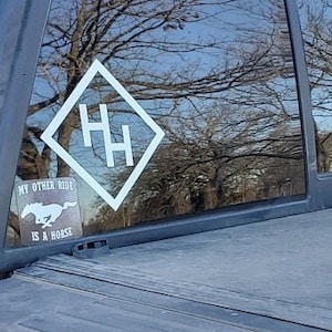 May include: A black truck with a black tonneau cover and a white sticker on the back window. The sticker says "My other ride is a horse" and has a white horse head logo and a white diamond with the letters "HH" inside.