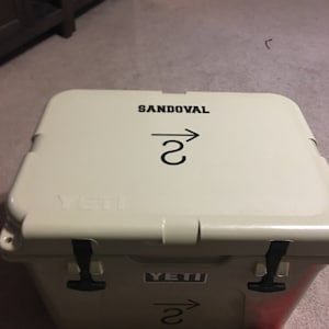 May include: A tan Yeti cooler with black handles and a black sticker that says "YETI". The cooler has a black sticker on the top that says "SANDOVAL" with an arrow pointing to the right and the number "2" underneath.