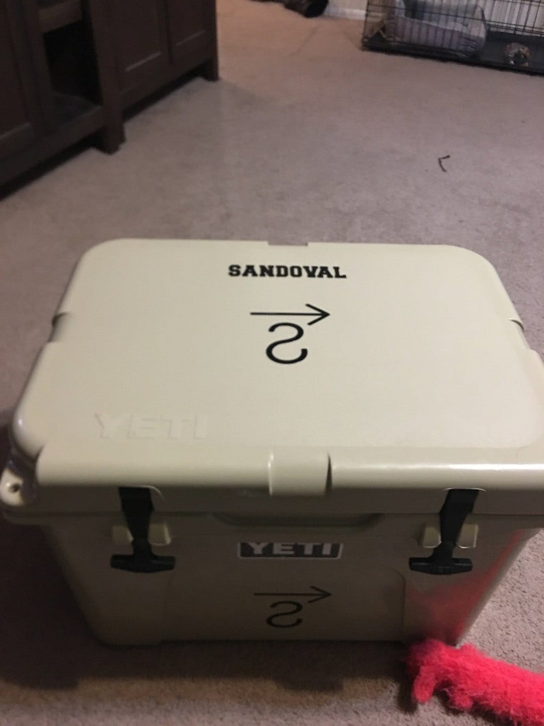 May include: A tan Yeti cooler with black handles and a black sticker that says "YETI". The cooler has a black sticker on the top that says "SANDOVAL" with an arrow pointing to the right and the number "2" underneath.