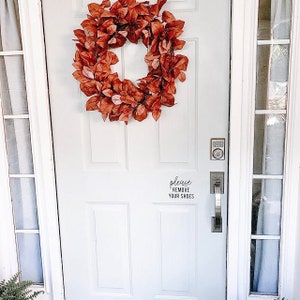 May include: A white front door with a wreath of faux fall leaves and a sign that reads "please remove your shoes".