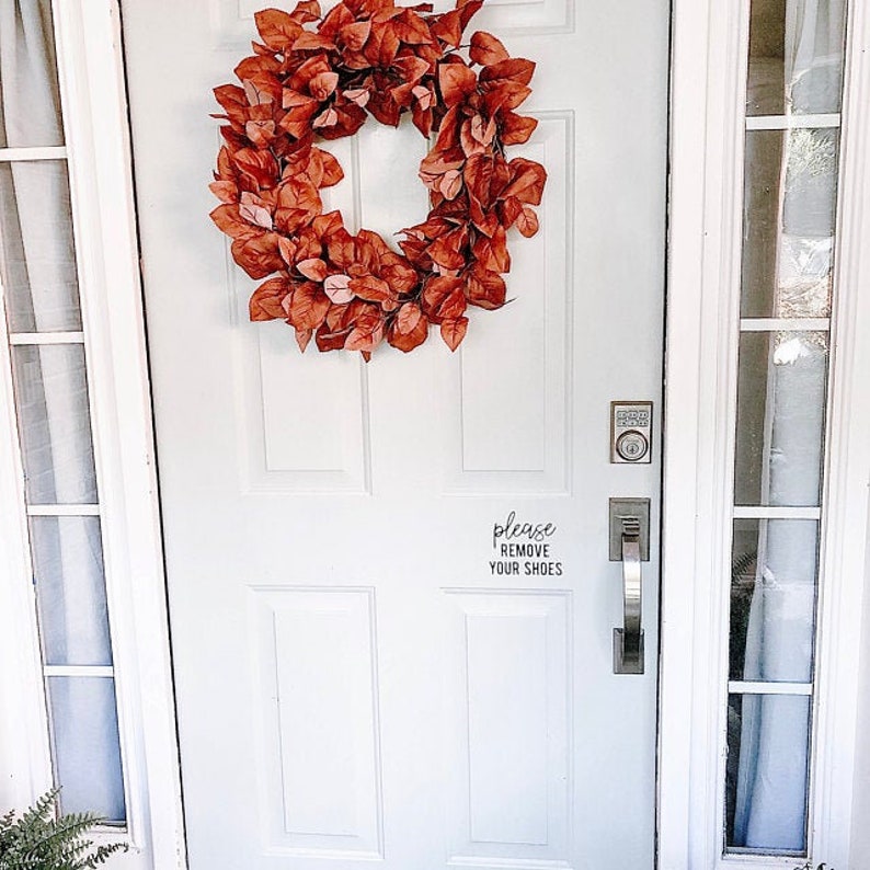 May include: A white front door with a wreath of faux fall leaves and a sign that reads "please remove your shoes".