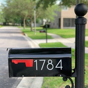 May include: Black mailbox with a red flag and white numbers "1784" on the front. The mailbox is mounted on a black post with a black ball finial.