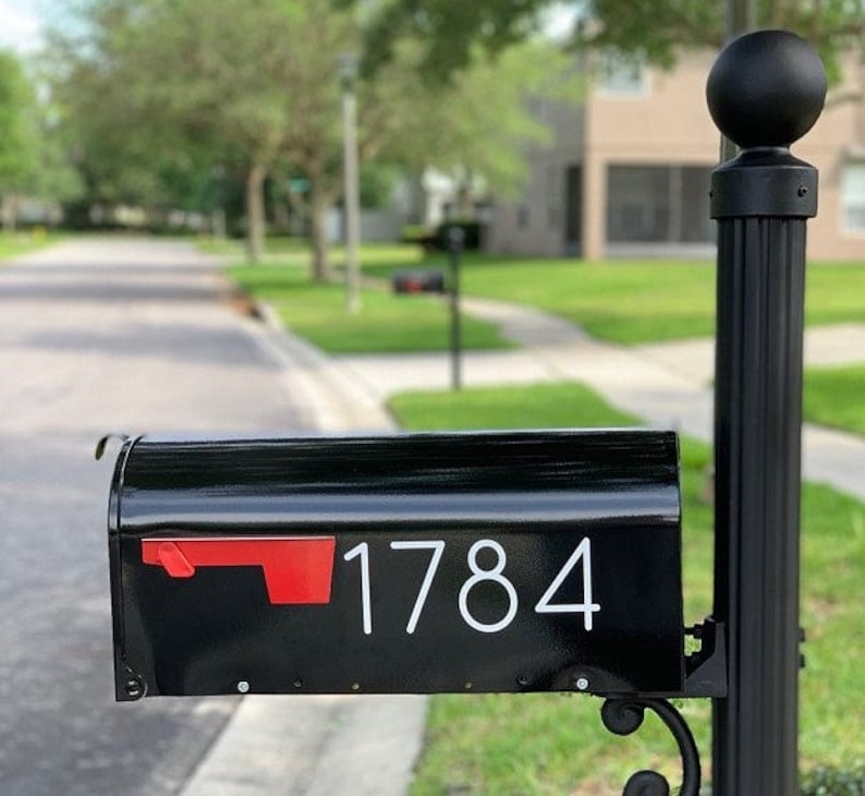 May include: Black mailbox with a red flag and white numbers "1784" on the front. The mailbox is mounted on a black post with a black ball finial.