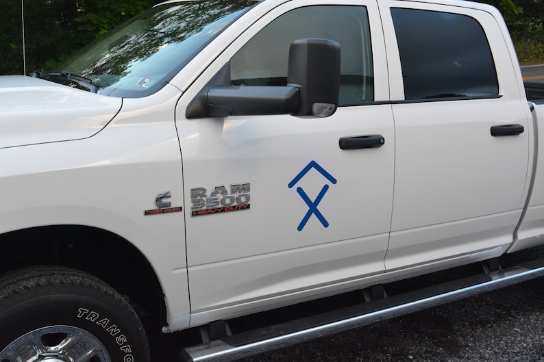 May include: White pickup truck with a blue logo on the side. The logo is a stylized arrow pointing upwards with an X underneath. The truck has a "RAM 3500 HEAVY DUTY" emblem on the front fender.