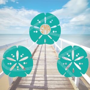 May include: Three teal blue sand dollar shapes on a wooden boardwalk with a blue sky and ocean in the background. The center sand dollar has the letters "SMA" in a circle in the center.