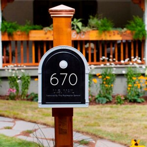 May include: A silver mailbox with a black door and a white number "670" on it. The mailbox is mounted on a wooden post.