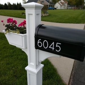 May include: A white mailbox post with a black mailbox. The mailbox has a white number "6045" on the front. There is a white planter box on the post with pink flowers.