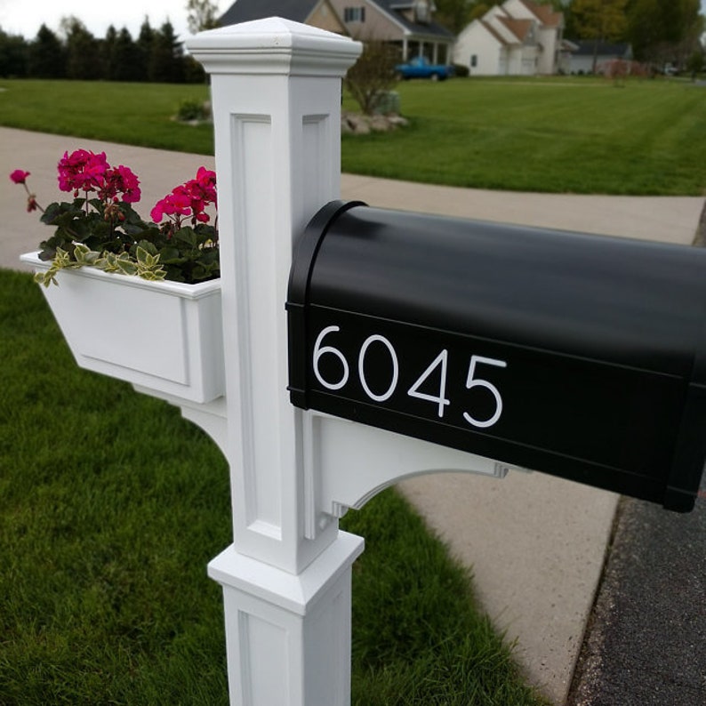 May include: A white mailbox post with a black mailbox. The mailbox has a white number "6045" on the front. There is a white planter box on the post with pink flowers.