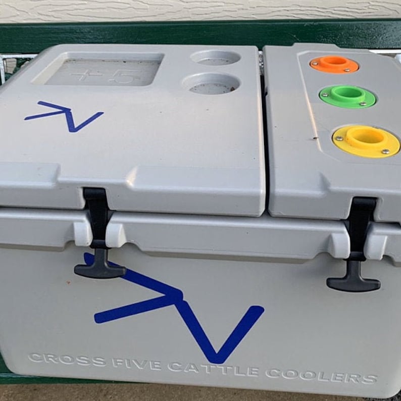 May include: A gray cooler with a blue logo that says "Cross Five Cattle Coolers". The cooler has a lid with a built-in table and cup holders. The lid also has three orange, green, and yellow colored cup holders.