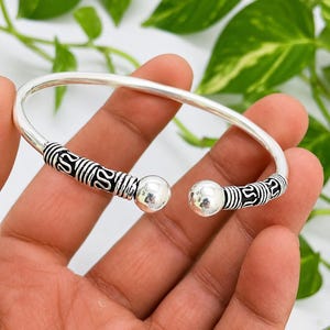 May include: A silver-colored bangle bracelet with decorative black and silver accents. The bracelet has a rounded shape with a ball at each end. The design includes intricate patterns and is held by a hand against a backdrop of green leaves.