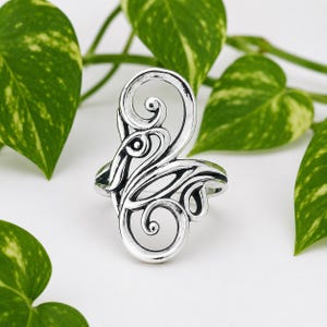 Può includere: Un anello color argento con un intricato design traforato di forme astratte e vorticose. L'anello è presentato su uno sfondo bianco, con foglie verdi sullo sfondo, creando un contrasto naturale.