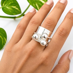 Puede incluir: Un anillo plateado con una banda ancha y tres bandas más pequeñas, cada una con una bola plateada. El anillo está en una mano, con una planta verde en el fondo. Una joya.