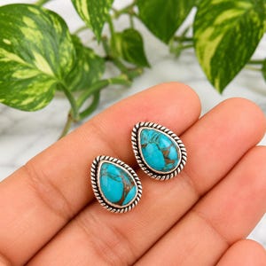 Op de afbeelding: Een paar druppelvormige turquoise oorstekers. De turkooizen stenen hebben bruine aderen en zijn gezet in een zilverkleurig metaal met een gedraaid touwontwerp rond de randen. De oorbellen worden op een hand getoond.