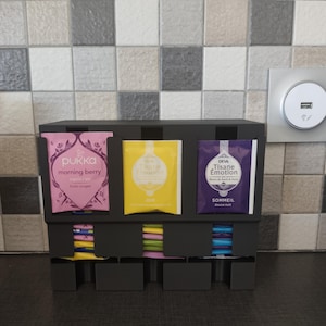 Könnte beinhalten: Ein schwarzer Teebeutel-Organizer mit drei vertikalen Fächern für einzelne Teebeutel. Die vorderen Beutel sind beschriftet mit Pukka Morning Berry in Pink, Deva Tisane Emotion Joie in Gelb und Deva Tisane Emotion Sommeil in Violett.