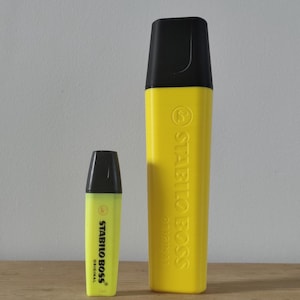 Peut inclure: Deux surligneurs Stabilo Boss Original jaunes avec des capuchons noirs. Le plus petit surligneur à gauche présente un corps jaune clair, tandis que le plus grand à droite est d'un jaune vif. Les deux portent le nom de la marque en noir.